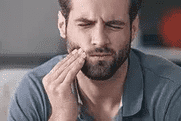 Bearded man touching his cheek in discomfort, suggesting dental pain or concern indoors.