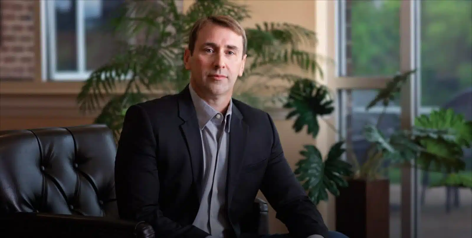 Professional man in dark blazer seated in modern office with plants and windows behind him.