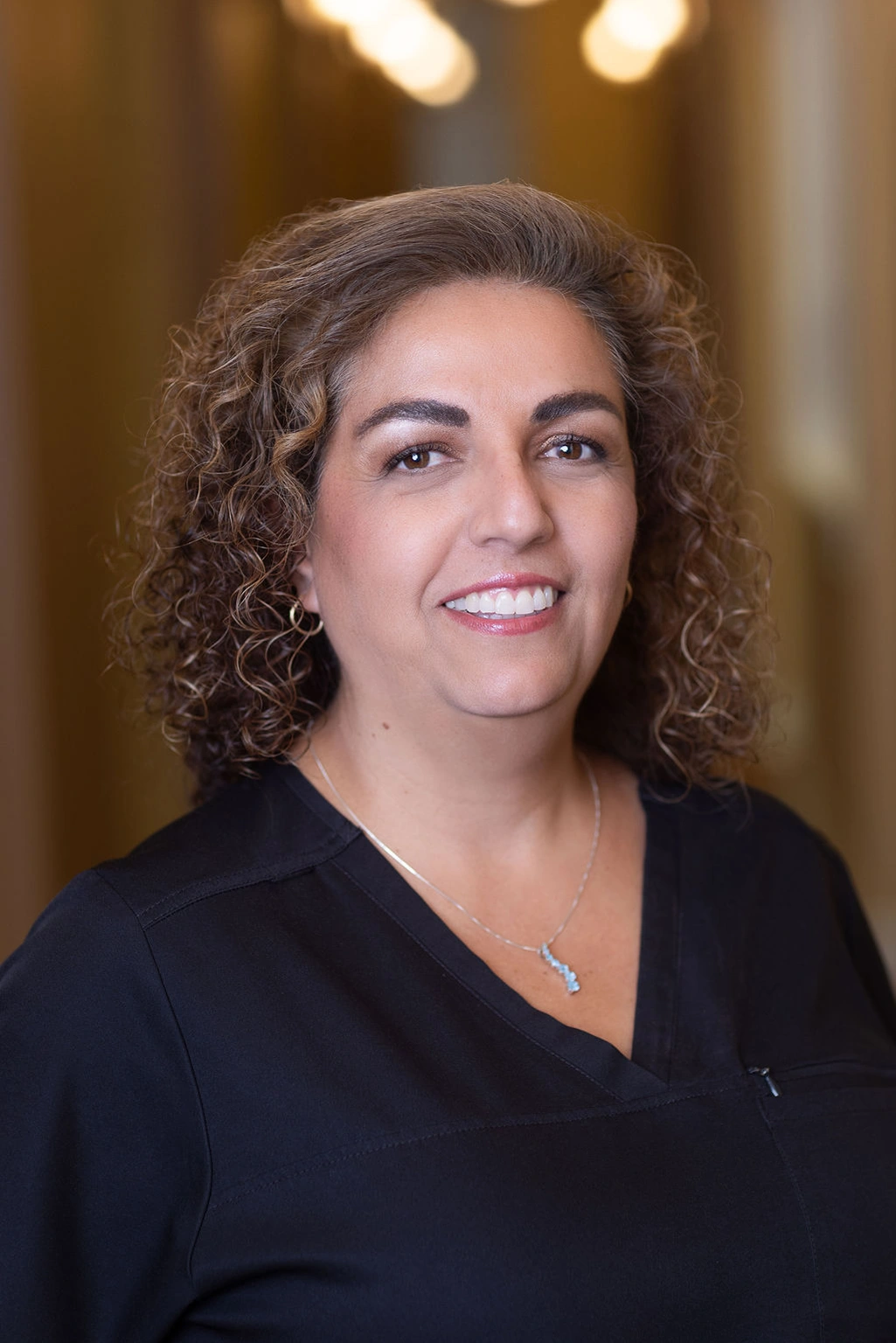 Woman with curly brown hair smiling in professional portrait, wearing black top, indoors.